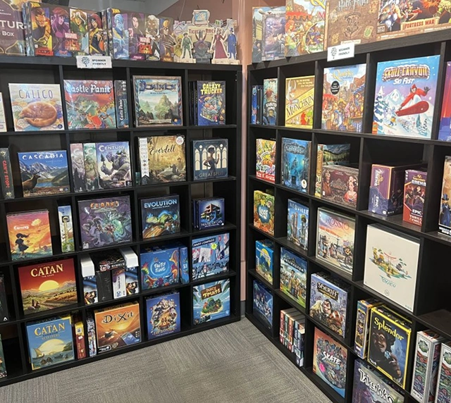 Planken vol met een verscheidenheid aan kleurrijke bordspellen, elk met uniek artwork. De scène straalt een levendige en uitnodigende hobbywinkel uit.
